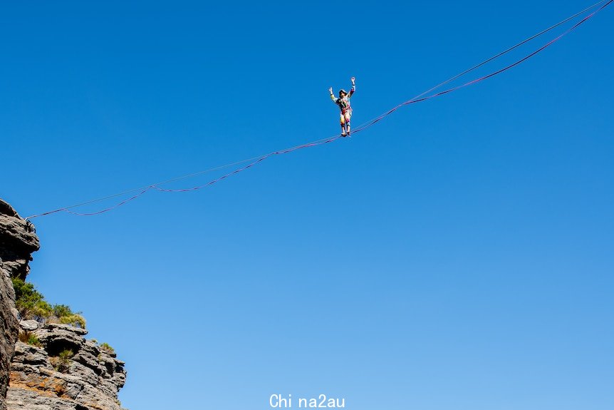 A figure high up on a wire, with just one cliff face visible under a blue sky.