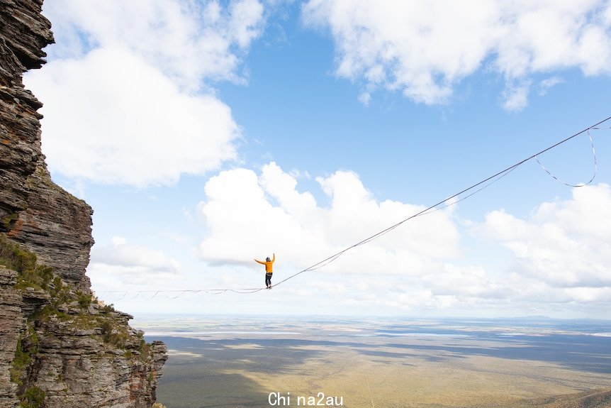 A figure suspended high above the valley between two cliff faces on a slackline, sunny skies.