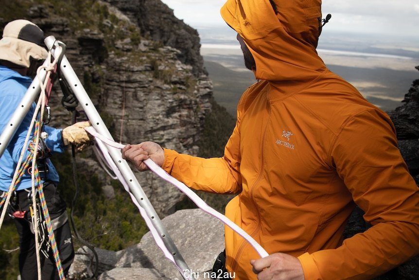 Man in orange jackets holds on a war in front of a cliff.