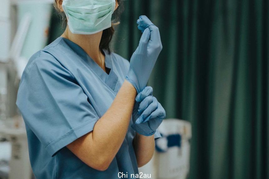 A masked nurse adjusts their blue plastic glove in an operation room.