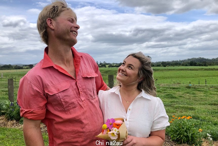 A small woman smiles up at her laughing husband, holding a bouquet of flowers on their farm.