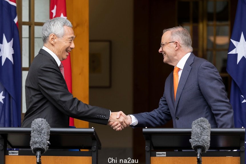 Two men in suits shake hands while standing in front of Australian flags.