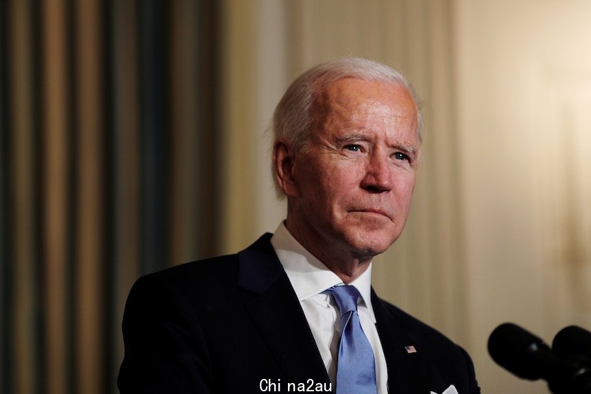 President Biden stands in front of a microphone wearing a black suit and blue tie with an American flag pinned to his lapel