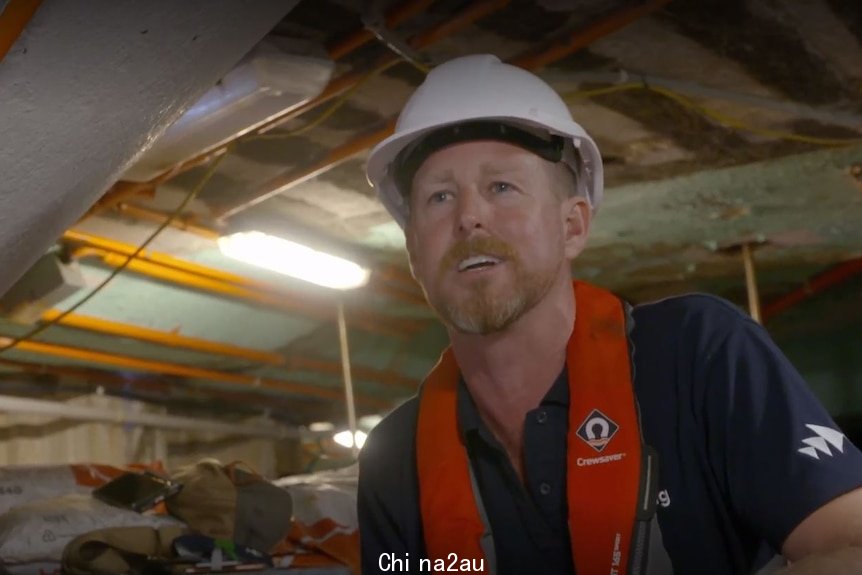 White man wearing construction hard hat and red safety vest speaks to camera from inside an underwater workshop.