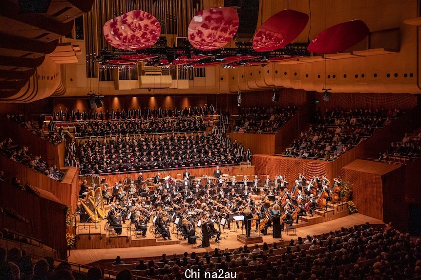 A large orchestra and choir performs on the Sydney Opera House stage. Magenta acoustic reflectors are suspended from ceiling. 