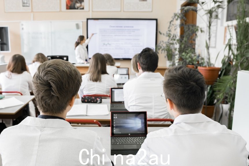 一排排学生穿着有领制服，坐在桌前使用小型笔记本电脑，一位老师则指着一块白板。