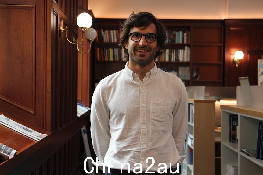 A man stands in an office. There are bookshelves and newspapers in the background.