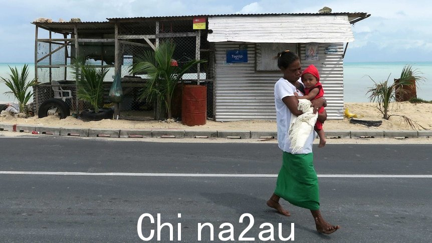 A mother carries a baby in front of a dilapidated shack