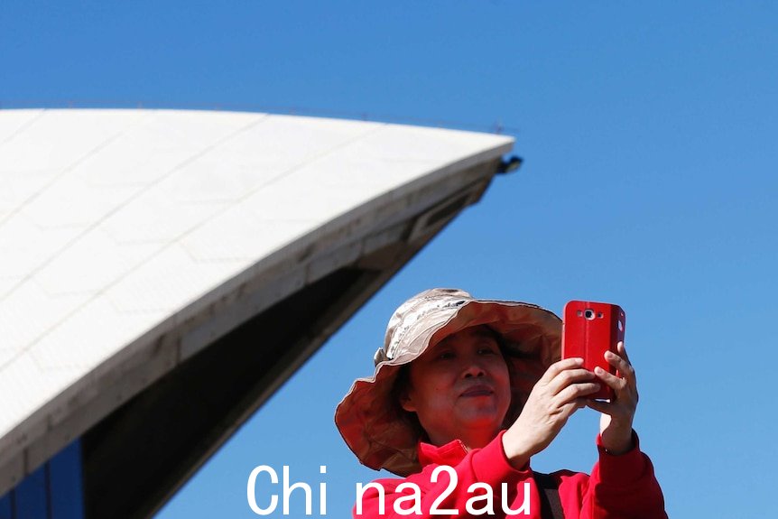 On a bright blue day, you see an Asian woman in bright pink take a selfie in front of one of the Sydney Opera House sails.