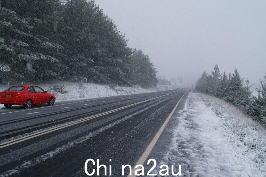 A red car drives down a straight road with snow falling all around it