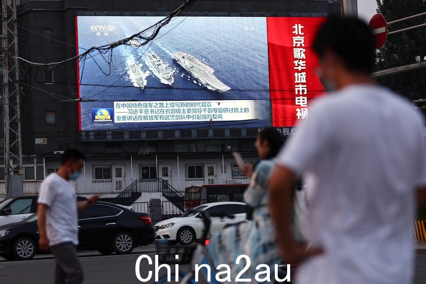 People in the street look up at a screen showing a news bulletin with images of Chinese warships