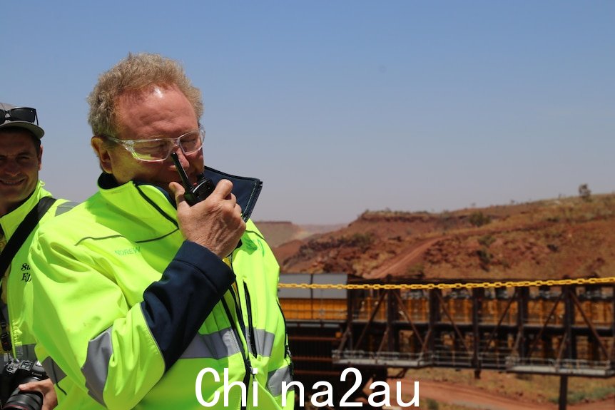 A man with a bright yellow jacket holds a walkie talkie in front of his mouth. He's standing at a mine site.