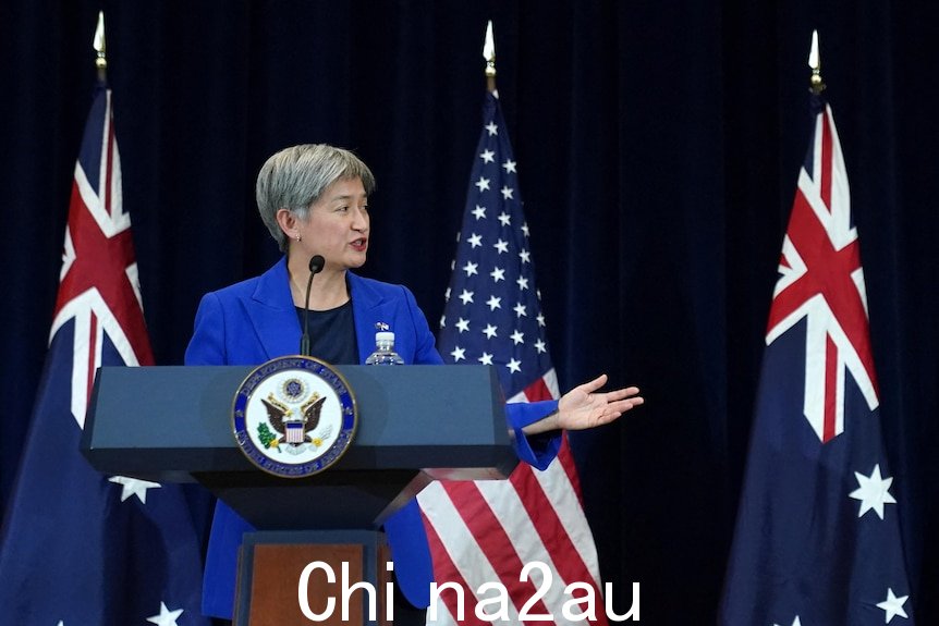 一位女士向一位男士做手势，与她一起站在澳大利亚和美国国旗前的舞台上。