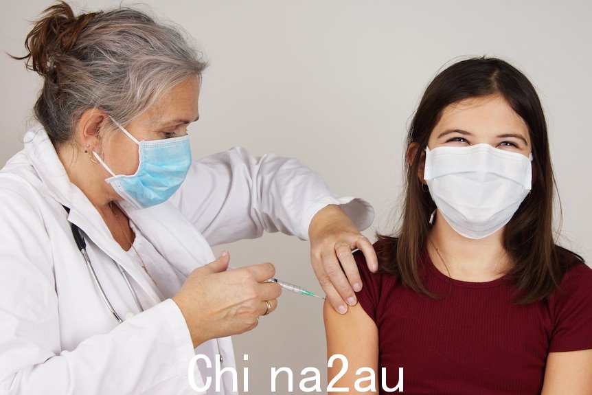 一名身穿白大衣、戴着蓝色口罩的女士为一名十几岁的女孩接种疫苗，她的面具下似乎在微笑。