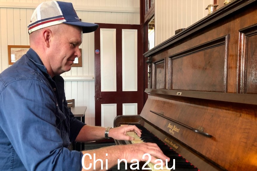 一个戴着帽子、身穿蓝色工作衬衫的男人坐着玩钢琴。” width=
