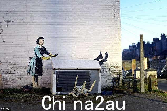 这幅画,许多人推测是在肯特郡马盖特的格罗夫纳广场附近,似乎描绘了一位 1950 年代的家庭主妇,她戴着蓝色的围巾和黄色的洗碗手套,将她的伴侣推入胸膛冰箱。” class=