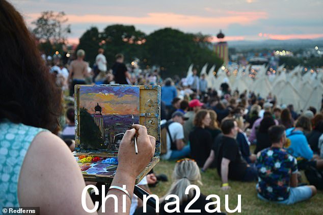 一位艺术家在 2023 年格拉斯顿伯里音乐节第一天的日落期间画了一幅画,人群坐着欣赏这一活动