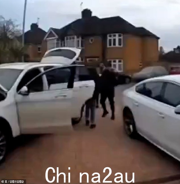 两名女性带着他参观那辆白色奔驰,一名男子双臂交叉站在后面,这辆车停在铺好的车道上,车门和引擎盖都打开