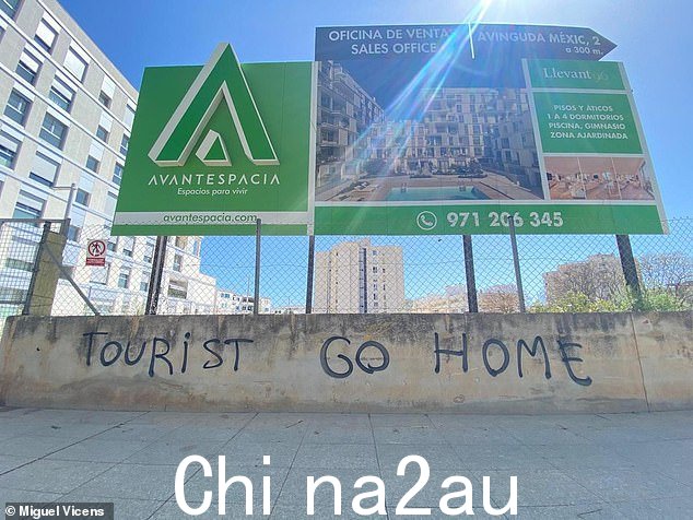 马略卡岛 Nou Llevant 的房地产促销广告牌下方的墙上用英文潦草地写着“回家旅游”字样,过去几年外国买家大量涌入该社区”class =