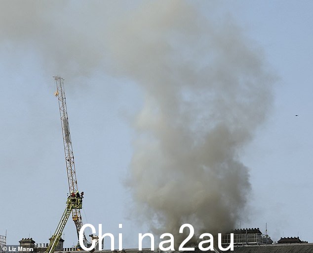 在首次报告火灾两小时后,烟雾继续从建筑物中冒出。可以看到一架无人机(右)正在勘察事故现场,消防员则从梯子上观看” class=