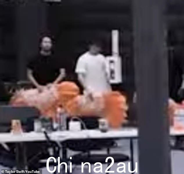 穿着白色T恤和黑色裤子,他闲散地站在一名男性伴舞旁边,挥舞着斯威夫特表演中使用的橙色羽毛扇道具